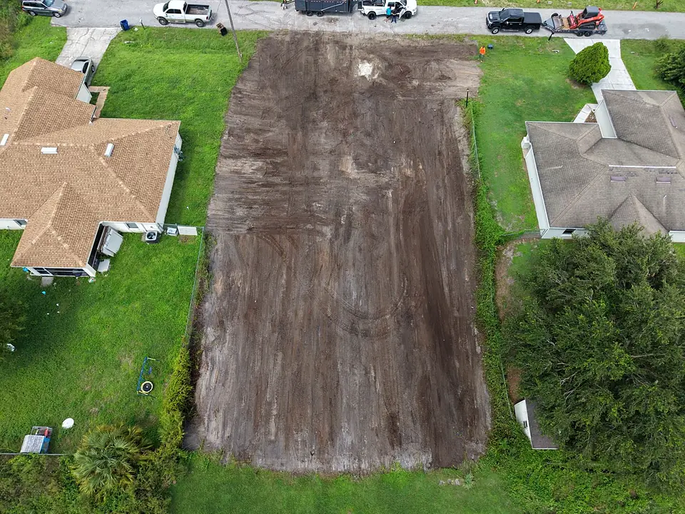 Aerial top-down view of a long cleared dirt lot between houses with trucks on the road