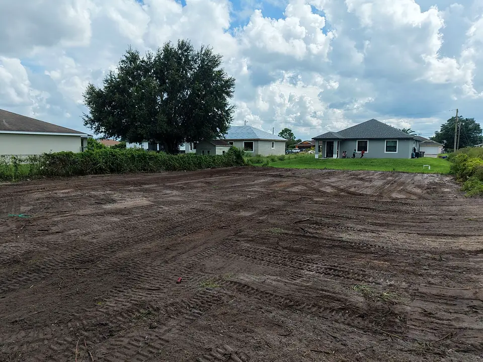 Wide cleared dirt lot with tire tracks, a large oak tree and houses beyond