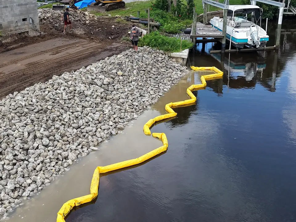 Aerial view of a long rip-rap canal bank with yellow turbidity barrier and a boat lift