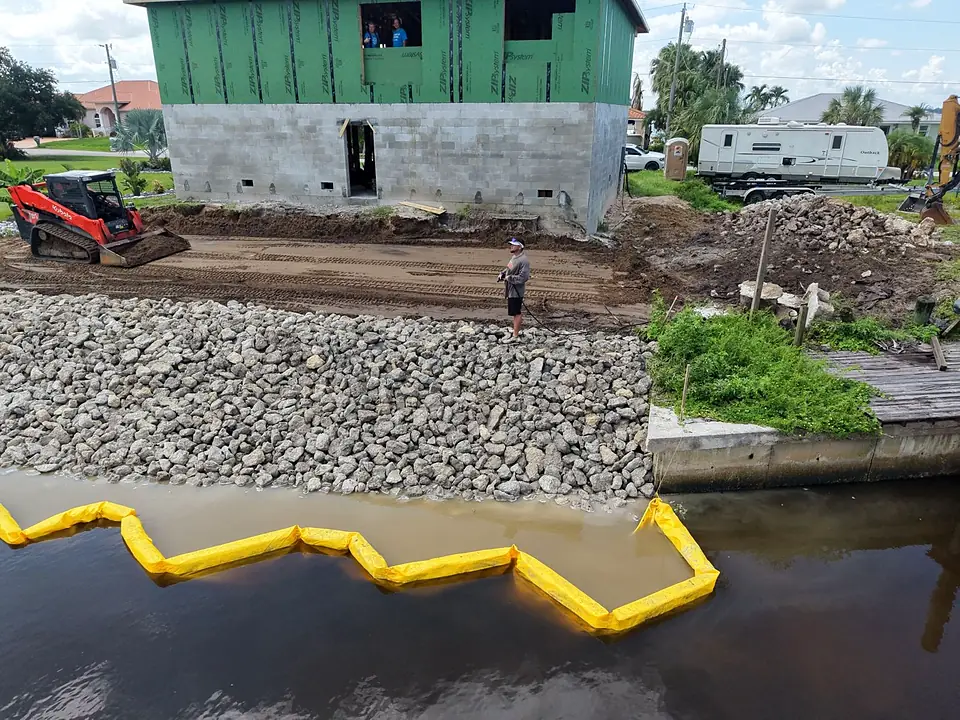 Aerial view of a rip-rap canal bank with yellow turbidity barrier and a track loader