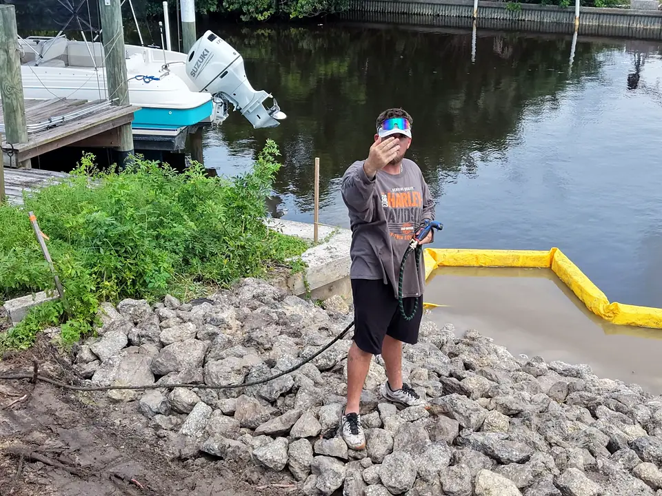 Worker holding a hose stands on rip-rap stone at a canal edge near a boat